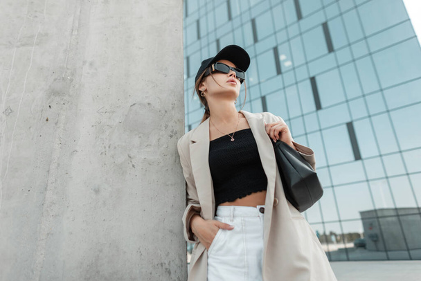 Fashionable urban beautiful woman model with a cool cap and black sunglasses with casual fashion clothes with a blazer and a top with a bag stands near a gray concrete wall in the city - Foto, Imagen