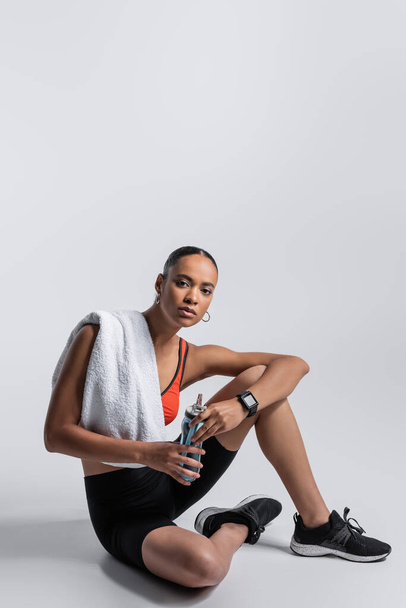 african american woman in red sports bra and bike shorts sitting with sports bottle and towel on grey  - Foto, immagini