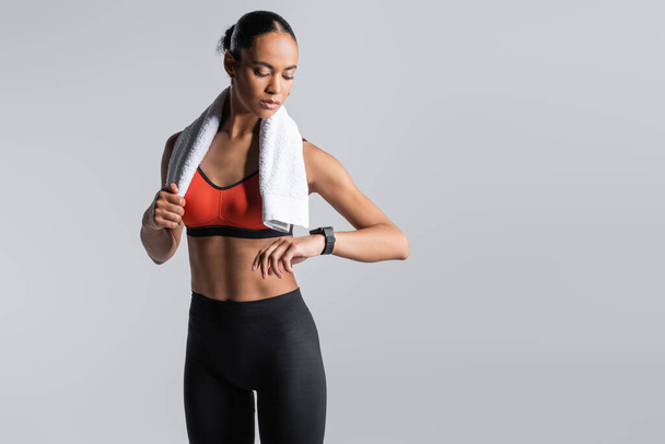 brunette african american sportswoman in red sports bra standing with towel and looking at fitness tracker isolated on grey  - Photo, Image