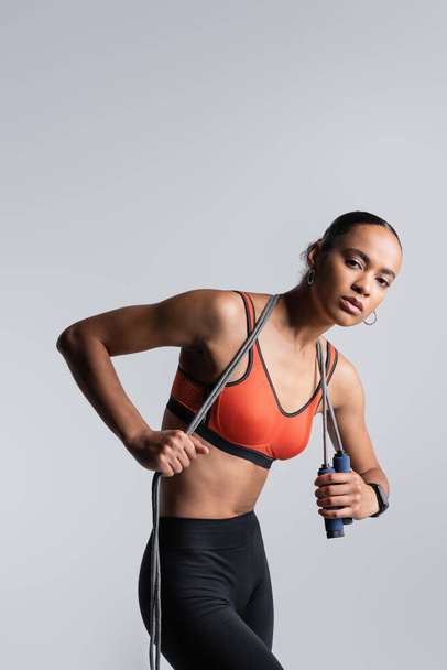 young african american sportswoman in red sports bra and bike shorts holding skipping rope isolated on grey  - Photo, image