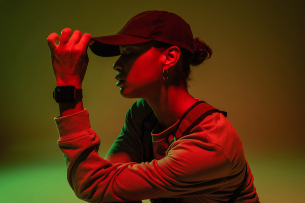 side view of african american woman in sweatshirt adjusting baseball cap on green with red light  - Foto, Imagen
