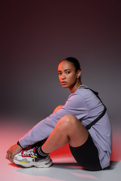 full length of young african american woman in bike shorts and sweatshirt sitting on grey with red light  - Fotografie, Obrázek