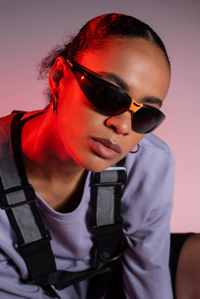 african american woman in sweatshirt and stylish sunglasses on grey with red light  - Foto, Imagen