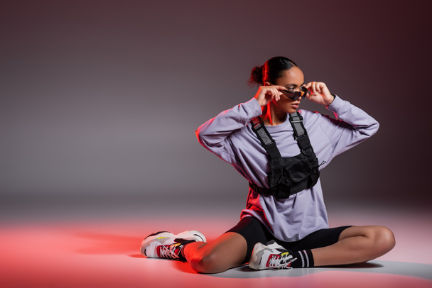full length of african american woman in bike shorts and sweatshirt wearing sunglasses on grey background with red light  - Fotoğraf, Görsel