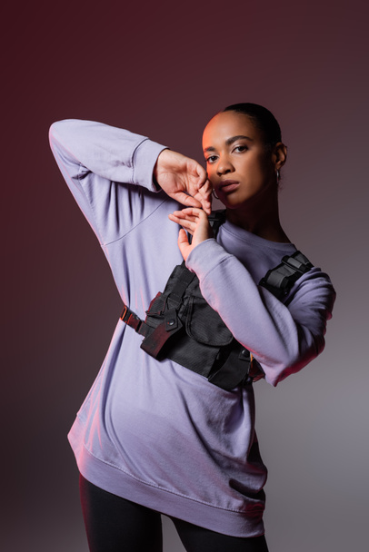 young african american woman in sweatshirt and vest looking at camera while posing on grey and purple  - Foto, Bild