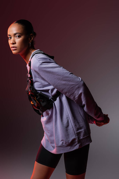 young african american woman in sweatshirt and vest looking at camera on grey and purple  - Foto, imagen