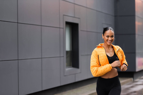 positive african american sportswoman in yellow puffer jacket jogging outside  - Φωτογραφία, εικόνα