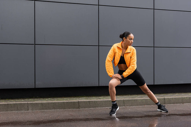 full length of brunette african american sportswoman in bike shorts and yellow puffer jacket exercising outside  - Фото, изображение