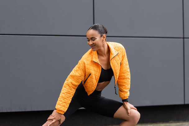 cheerful african american sportswoman in bike shorts and yellow puffer jacket exercising outside - Photo, image