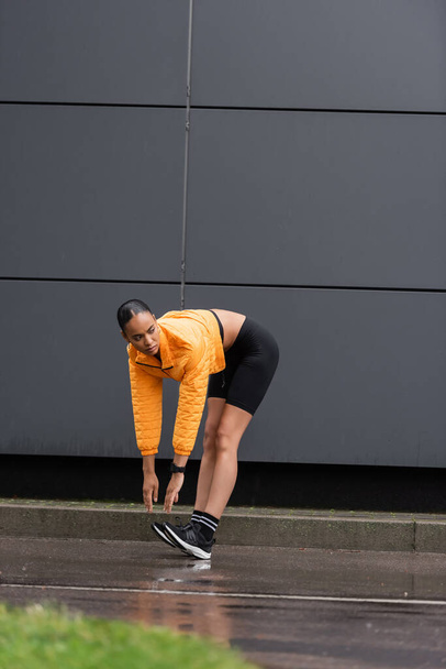 full length of african american sportswoman in bike shorts and yellow puffer jacket stretching near grey building  - Φωτογραφία, εικόνα