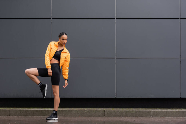 full length of brunette african american sportswoman in bike shorts and yellow puffer jacket posing outside  - 写真・画像