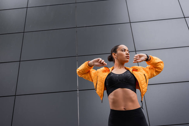 low angle view of brunette african american sportswoman in sports bra and yellow puffer jacket exercising outside  - 写真・画像