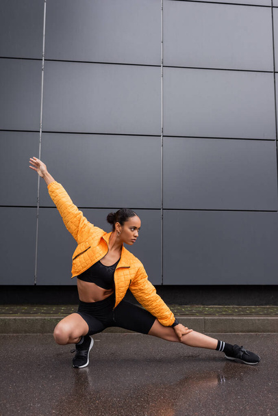 full length of stylish african american sportswoman in bike shorts and yellow puffer jacket stretching leg near grey building  - Foto, imagen