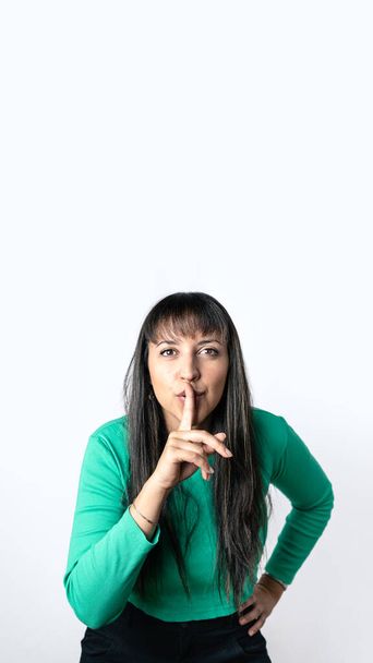 Brown hair woman with Hispanic features making silence gesture with her finger on lips over isolated white background,. Vertical photography with copy space above - Zdjęcie, obraz