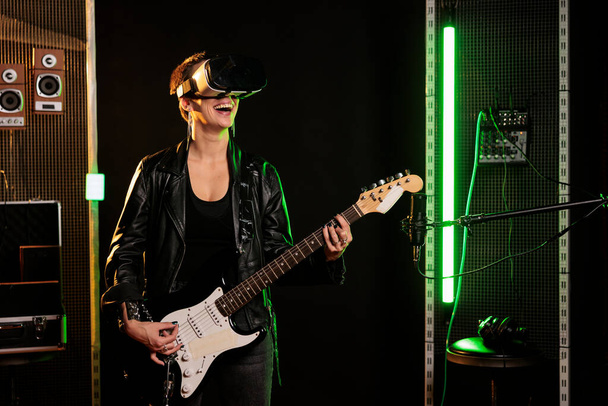 Guitarist with virtual reality headset enjoying rock concert simulation while playing heavy metal song at electric guitar in music studio. Woman musician performing grunge album using - Photo, image