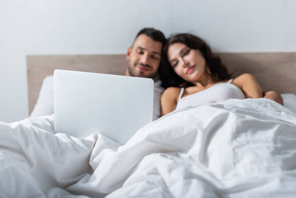 Laptop near blurred couple lying on bed in morning  - Fotó, kép