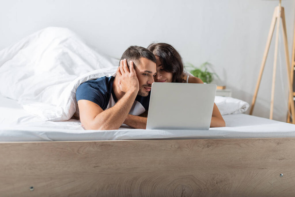 Молодой человек смотрит на ноутбук рядом с брюнеткой подруги на кровати  - Фото, изображение