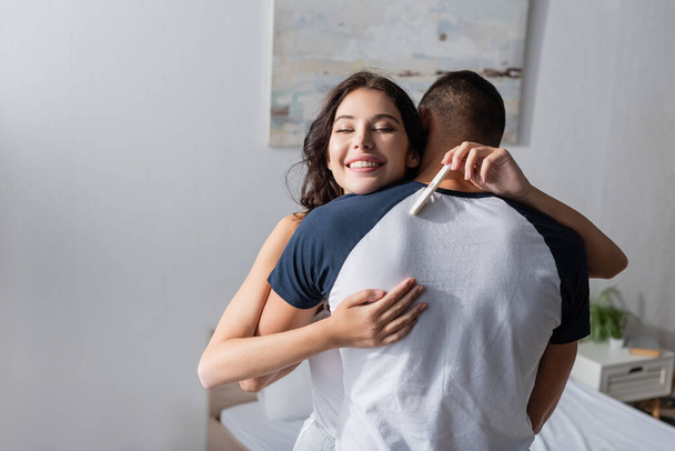 Happy woman holding pregnancy test and embracing boyfriend in bedroom  - Foto, Imagen