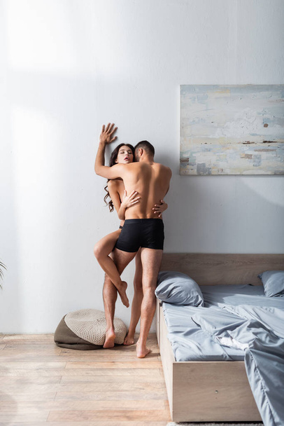 Мускулистый мужчина в трусах целует сексуальную девушку у кровати дома  - Фото, изображение