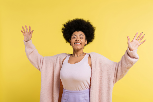 optimistic african american woman with trendy hairstyle smiling at camera and waving hands isolated on yellow - Foto, immagini