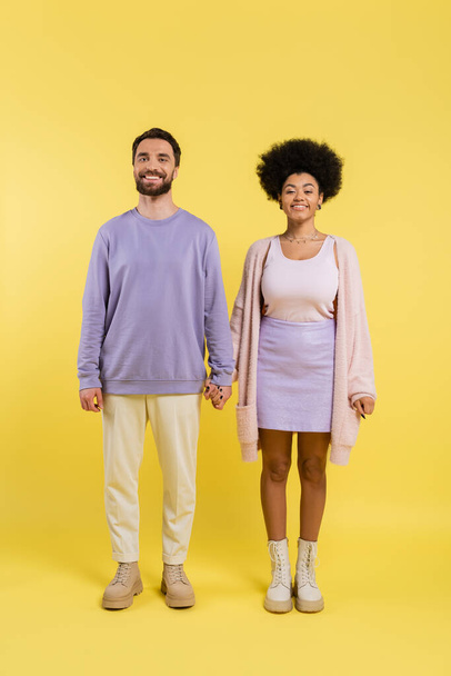 full length of trendy interracial couple holding hands and smiling at camera on yellow background - Foto, immagini