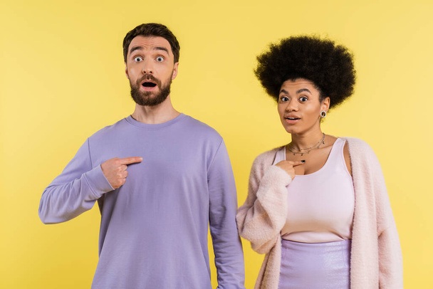 surprised interracial couple looking at camera and pointing with fingers at themselves isolated on yellow - Photo, image