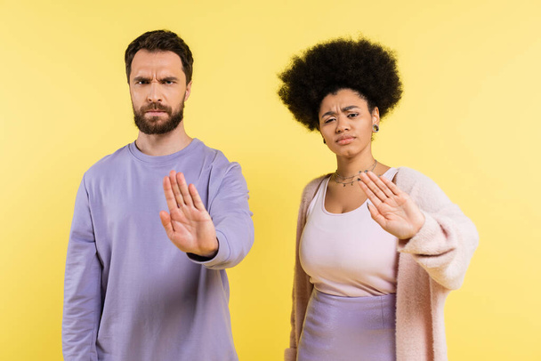 displeased and frowning multiethnic couple looking at camera and showing refuse gesture isolated on yellow - Zdjęcie, obraz