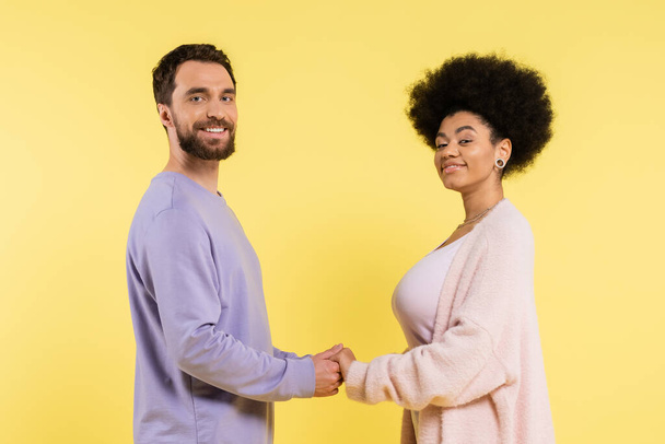 cheerful multiethnic couple holding hands and looking at camera while posing isolated on yellow - Foto, imagen