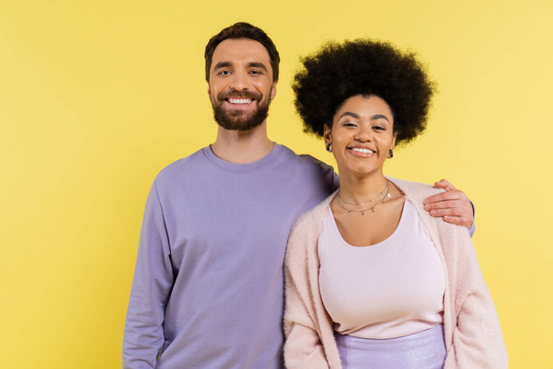 happy bearded man hugging shoulders of stylish african american woman while looking at camera isolated on yellow - Foto, imagen
