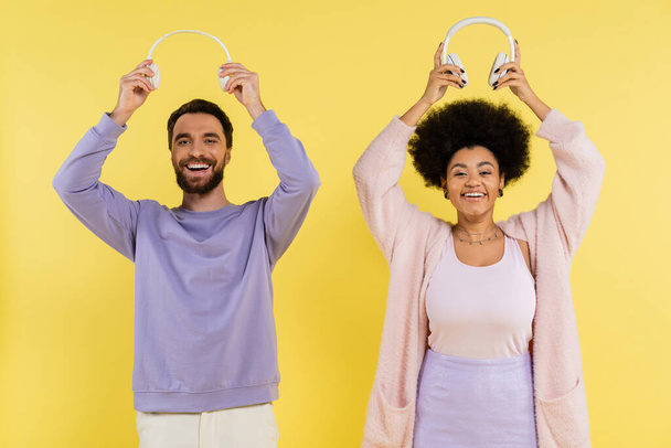 cheerful and trendy interracial couple holding wireless headphones above heads isolated on yellow - Zdjęcie, obraz