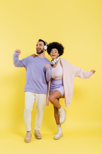 full length of excited and stylish multiethnic couple dancing in wireless headphones on yellow background - Фото, изображение