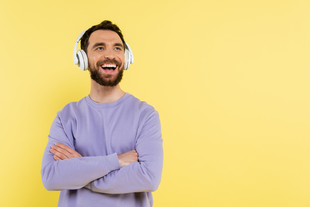 bearded man listening music in wireless headphones while standing with crossed arms and laughing isolated on yellow - Fotoğraf, Görsel