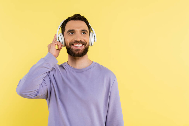happy bearded man adjusting wireless headphones while listening music isolated on yellow - Foto, afbeelding