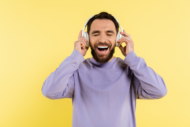excited bearded man adjusting wireless headphones and laughing with closed eyes isolated on yellow - Photo, image