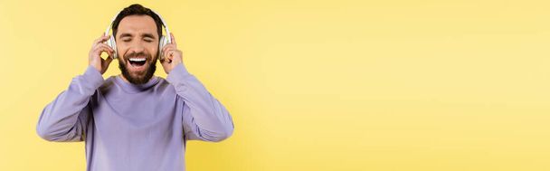cheerful man with closed eyes adjusting wireless headphones and laughing isolated on yellow, banner - Foto, afbeelding