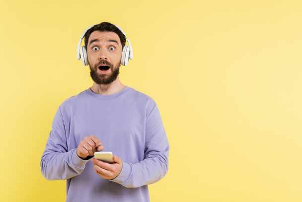 astonished man in wireless headphones holding mobile phone while looking at camera isolated on yellow - Foto, Imagem