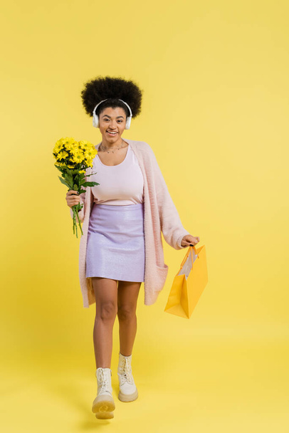 full length of smiling african american woman in wireless headphones walking with flowers and shopping bag on yellow background - Foto, Imagen