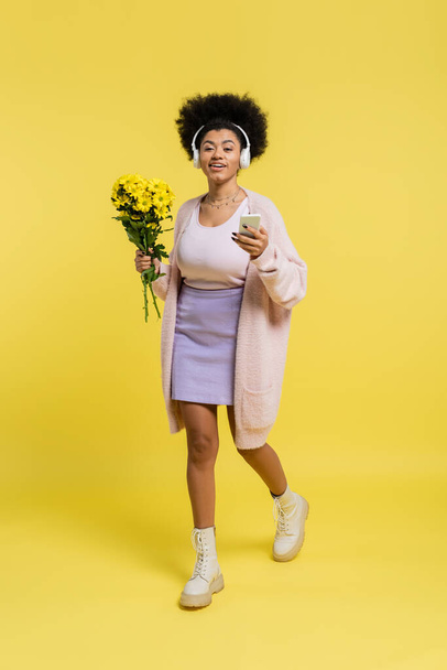 full length of happy african american woman in wireless headphones walking with bouquet and smartphone on yellow  - Фото, зображення