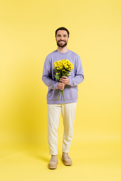 full length of bearded and trendy man holding bouquet and smiling at camera on yellow  - Photo, Image