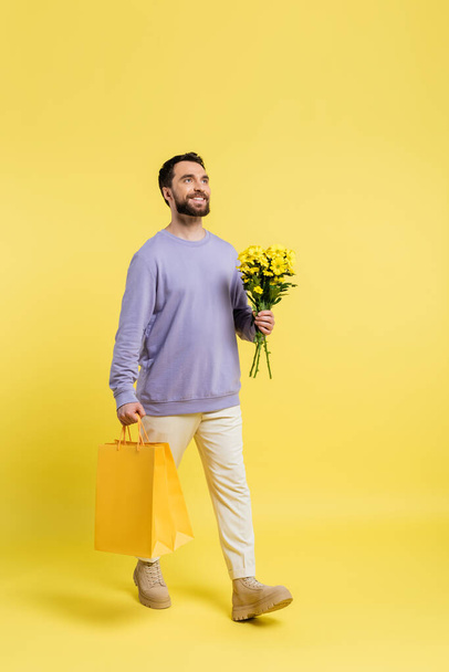 full length of happy and stylish man walking with shopping bags and flowers on yellow background - Foto, Imagen