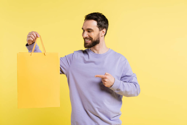 радостный человек в фиолетовый пуловер указывая пальцем на торговый мешок изолирован на желтый - Фото, изображение
