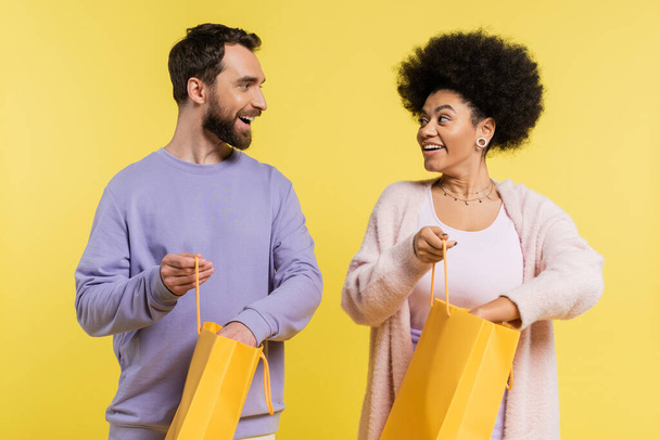 bearded man and curly african american woman holding shopping bags and smiling at each other isolated on yellow - Fotó, kép