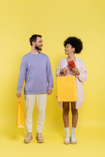 full length of trendy african american woman holding gift box and shopping bag near smiling bearded man on yellow  - Fotografie, Obrázek