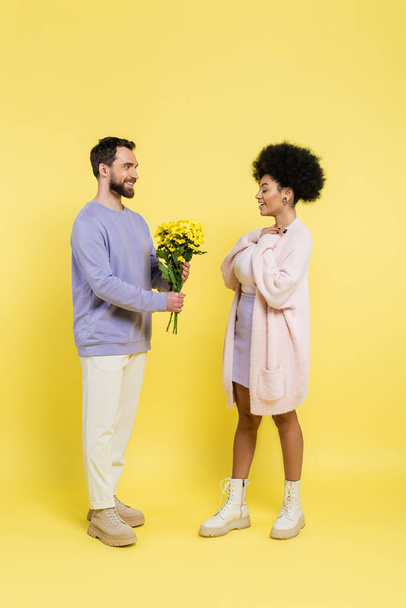 full length of smiling bearded man presenting flowers to pleased african american woman on yellow  - Foto, Imagem