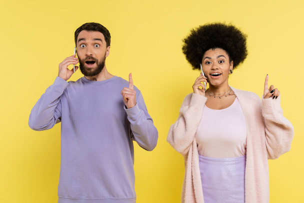 amazed multiethnic couple showing idea gesture while talking on smartphones isolated on yellow - Photo, image