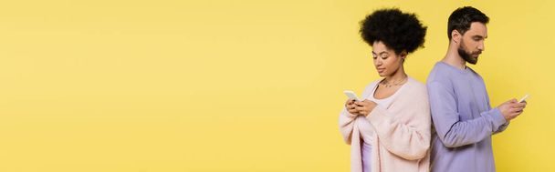 黄色のバナーに囲まれた携帯電話でおしゃべりしながら立っているスタイリッシュな異人種間のカップル - 写真・画像