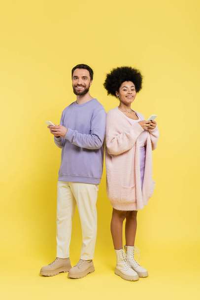 full length of happy and stylish multiethnic couple looking at camera while standing with cellphones on yellow  - Foto, Bild
