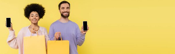 cheerful bearded man and curly african american woman with shopping bags showing smartphones with blank screen isolated on yellow, banner - Valokuva, kuva