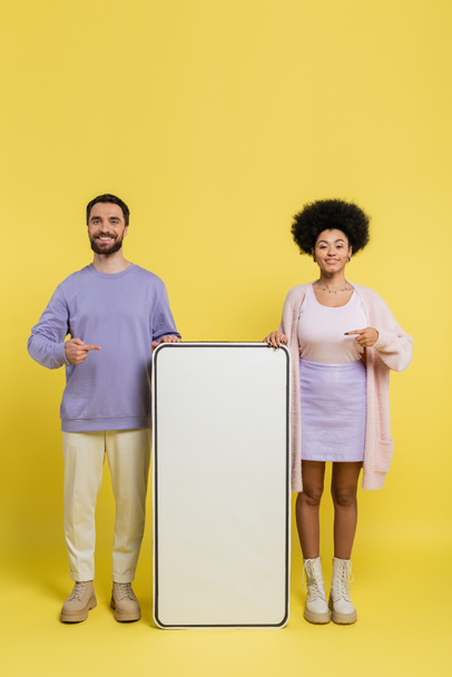 full length of cheerful multiethnic couple pointing at blank template of mobile phone on yellow background - Photo, image