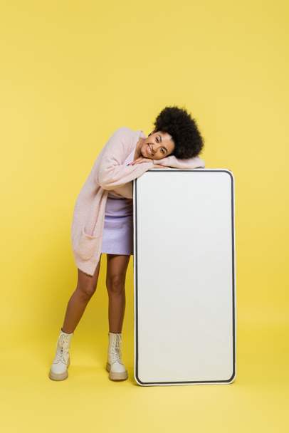 full length of happy and trendy african american woman posing with huge template of smartphone on yellow background - Foto, Imagem
